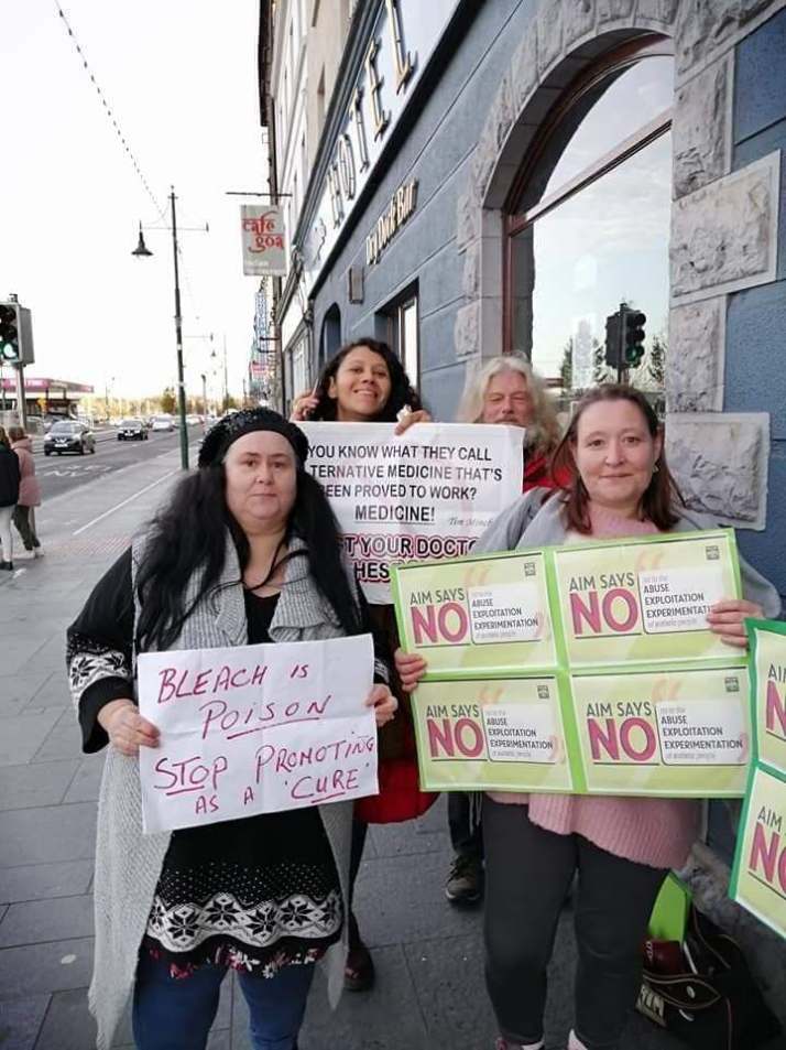 Three women stand outside a hotel, holding signs with messages including "AIM says NO to the Abuse / Exploitation / Experimentation of autistic people" and "Bleach is a Poison - Stop Promoting as a 'Cure'".