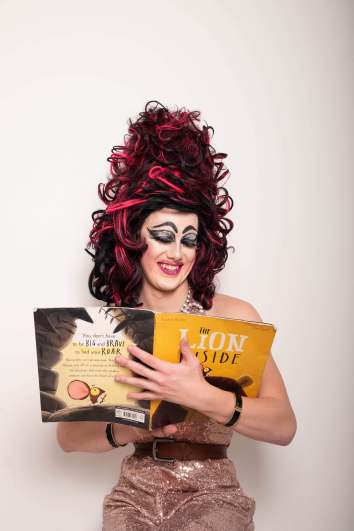 Aida H Dee, a drag queen with a red and black neon wig and dramatic eye makeup, smiles as she reads from a picture book entitled "The Lion Inside".